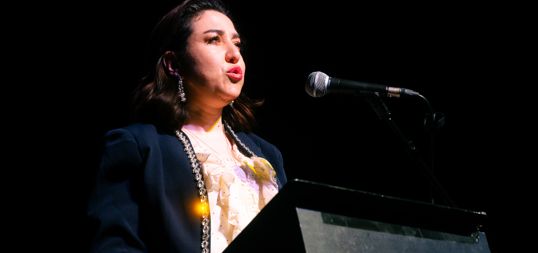 A woman with dark hair speaking in to a microphone in a dark room