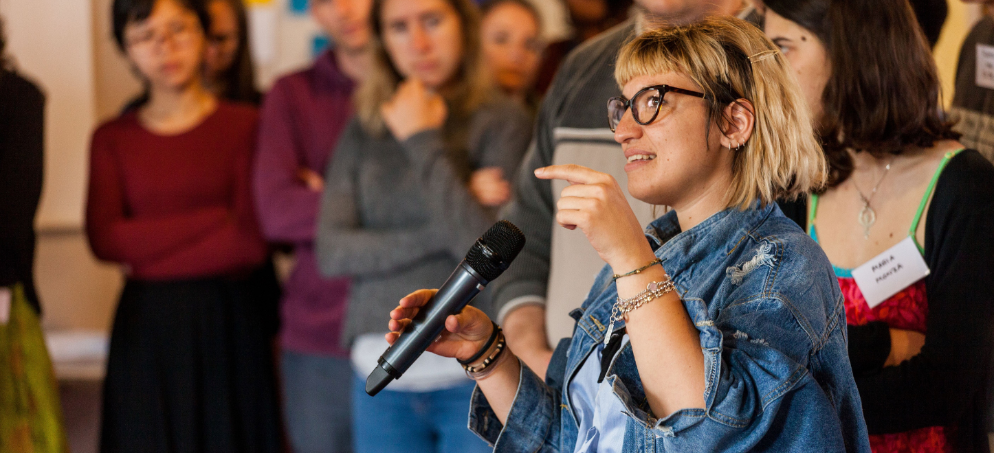 A woman with short blonde hair and glasses is speaking into a microphone.