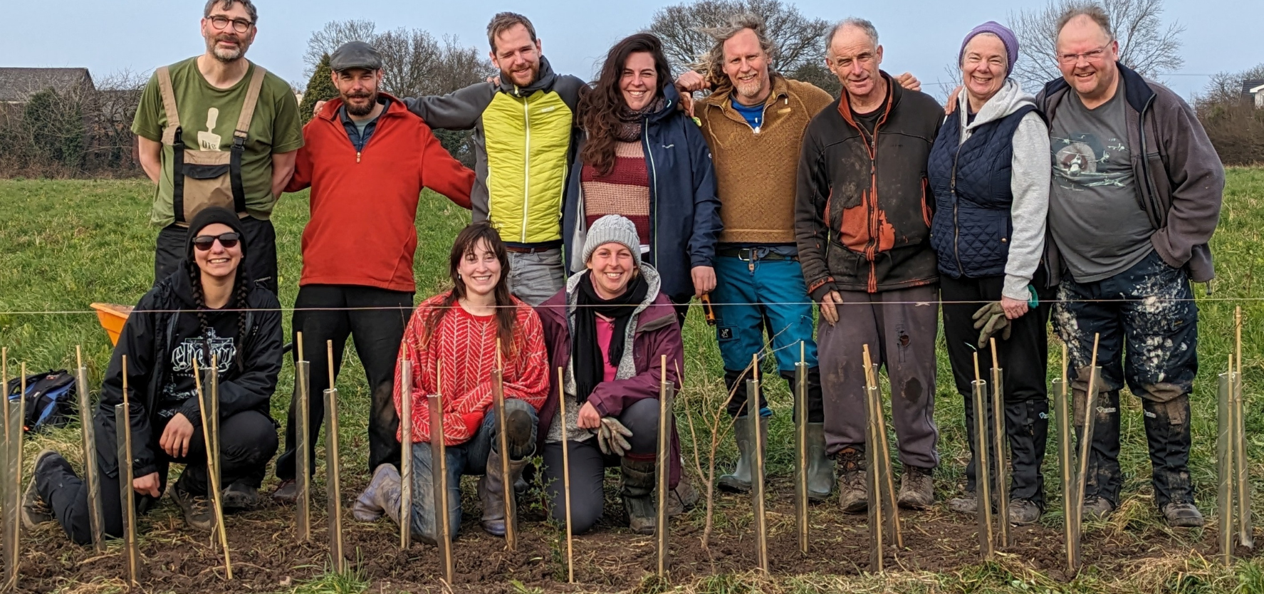 A group of people are standing with their arms around each other, three peple kneel in front. A row of sticks stand upright in the ground in front of them.