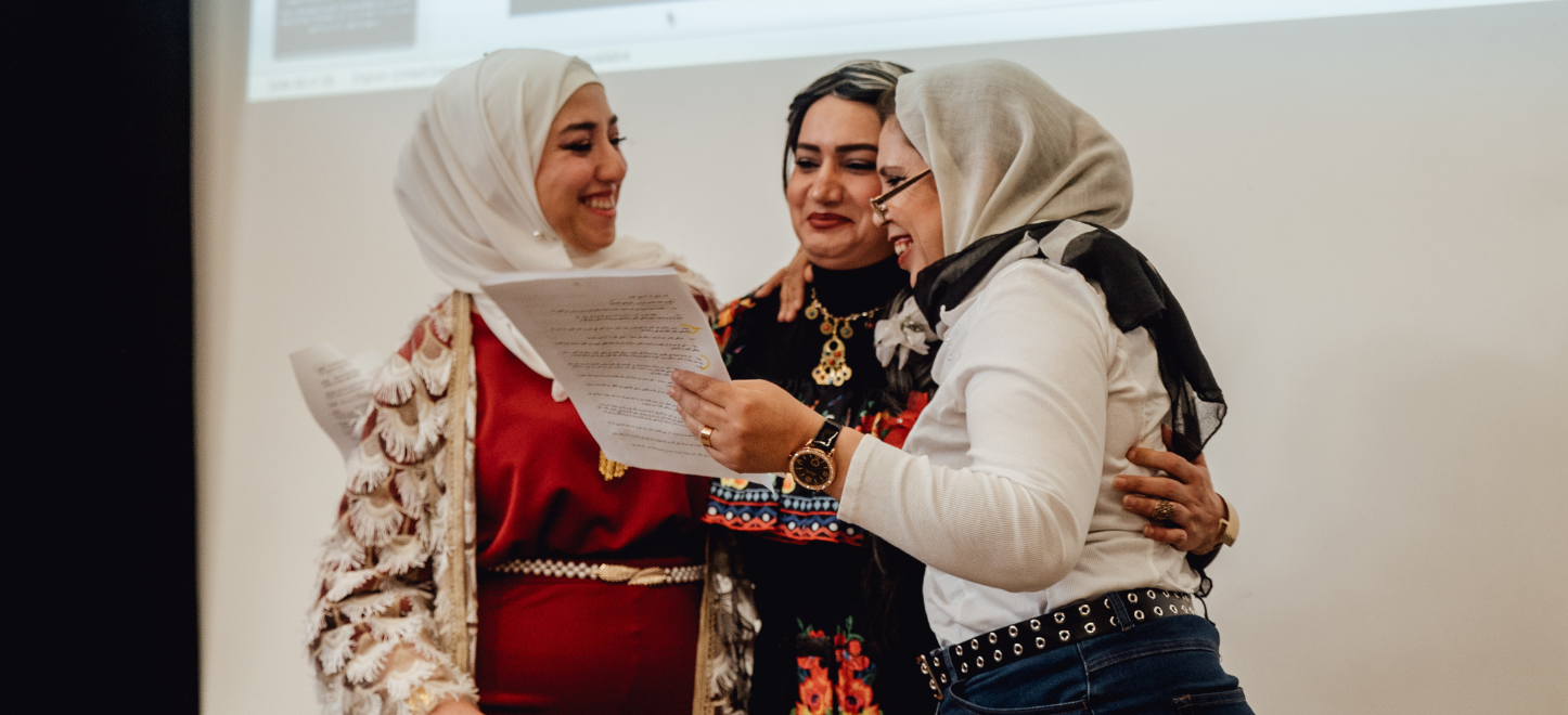 The picture shows three women stood close together, reading from a sheet of paper. Two of the women are wearing hijabs.