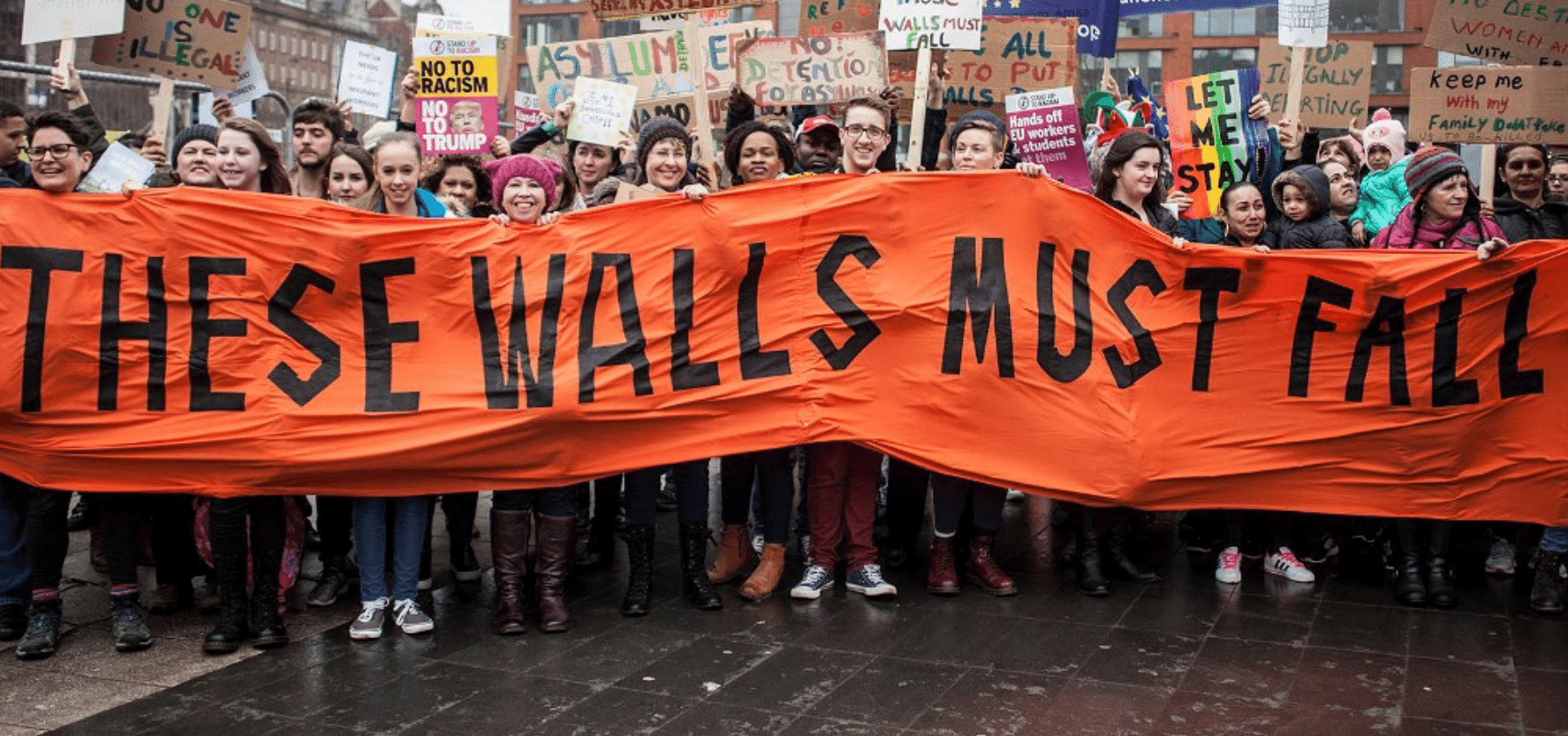 An image of a crowd of people holding an orange banner saying 'these walls must fall'. Picture: Right to Remain